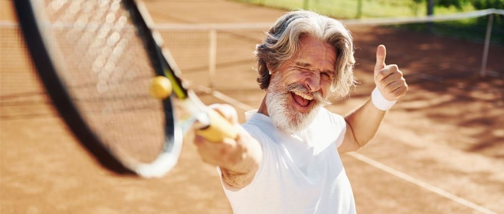 Älterer Mann mit grauem Haar spielt Tennis auf Sandplatz und zeigt Daumen hoch. Sportliche Stimmung im Freien.​​​​​​​​​​​​​​​​​​​​​​​​​​​​​​​​​​​