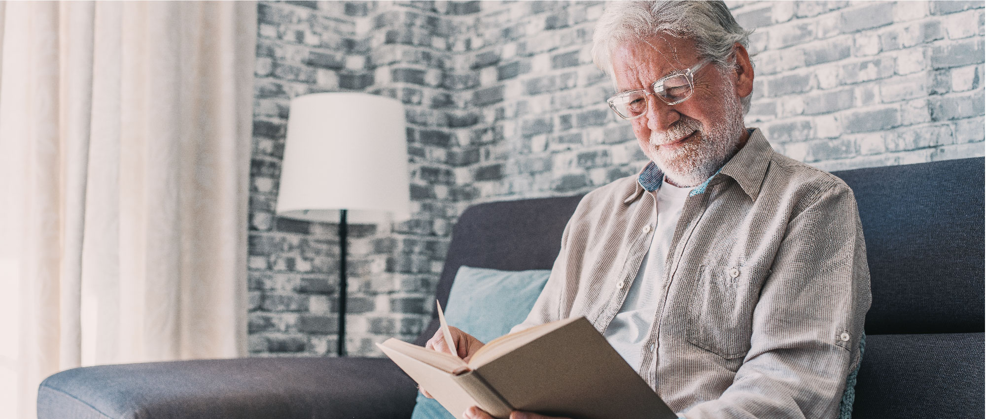 Älterer Mann mit Brille liest entspannt ein Buch auf dem Sofa vor Backsteinwand und Stehlampe im Wohnzimmer​​​​​​​​​​​​​​​​​​​​​​​​​​​​​​​​​​​​​​