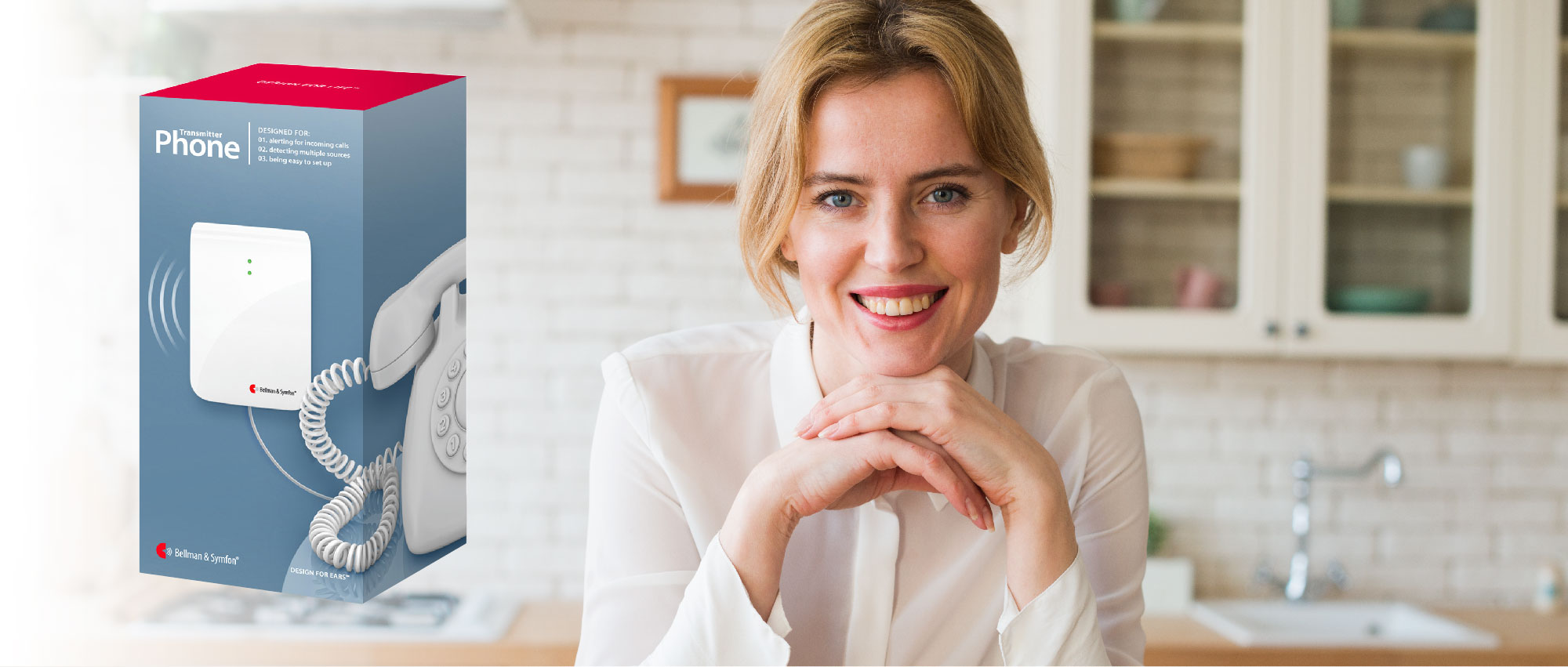 Lächelnde Frau in heller Küche neben Bellman & Symfon Telefon-Transmitter Verpackung für Hörunterstützung​​​​​​​​​​​​​​​​​​​​​​​​​​​​​​​​​​​​​​​​
