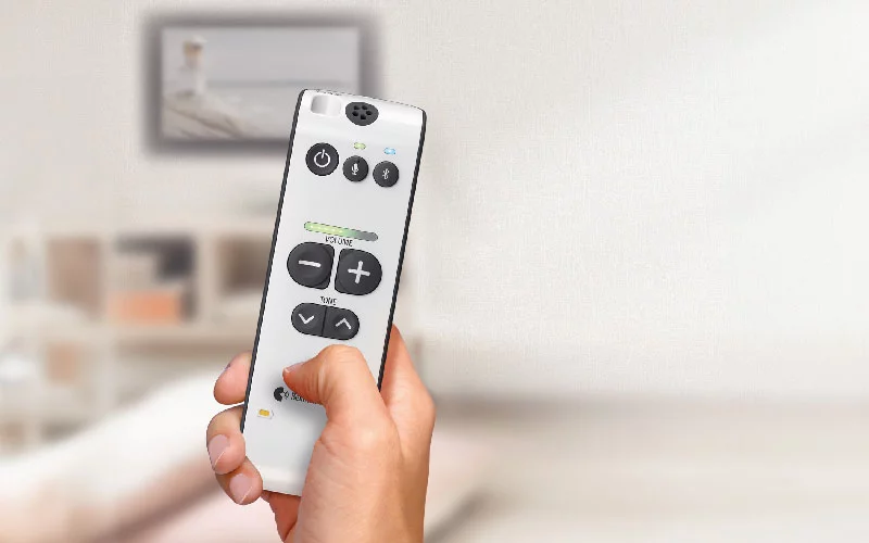 Hand hält Bellman Symfon Maxi Pro Fernbedienung vor unscharfem Wohnzimmerhintergrund mit Regal und Bild an der Wand​​​​​​​​​​​​​​​​​​​​​​​​​​​​​​