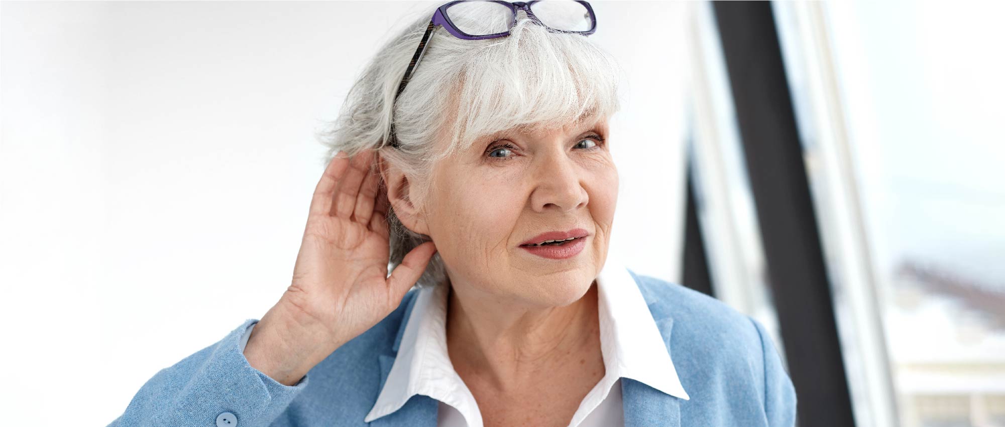 Ältere Frau mit grauen Haaren hält Hand ans Ohr und trägt keine Hörgeräte in heller Innenraumumgebung​​​​​​​​​​​​​​​​​​​​​​​​​​​​​​​​​​​​​​​​​​​​
