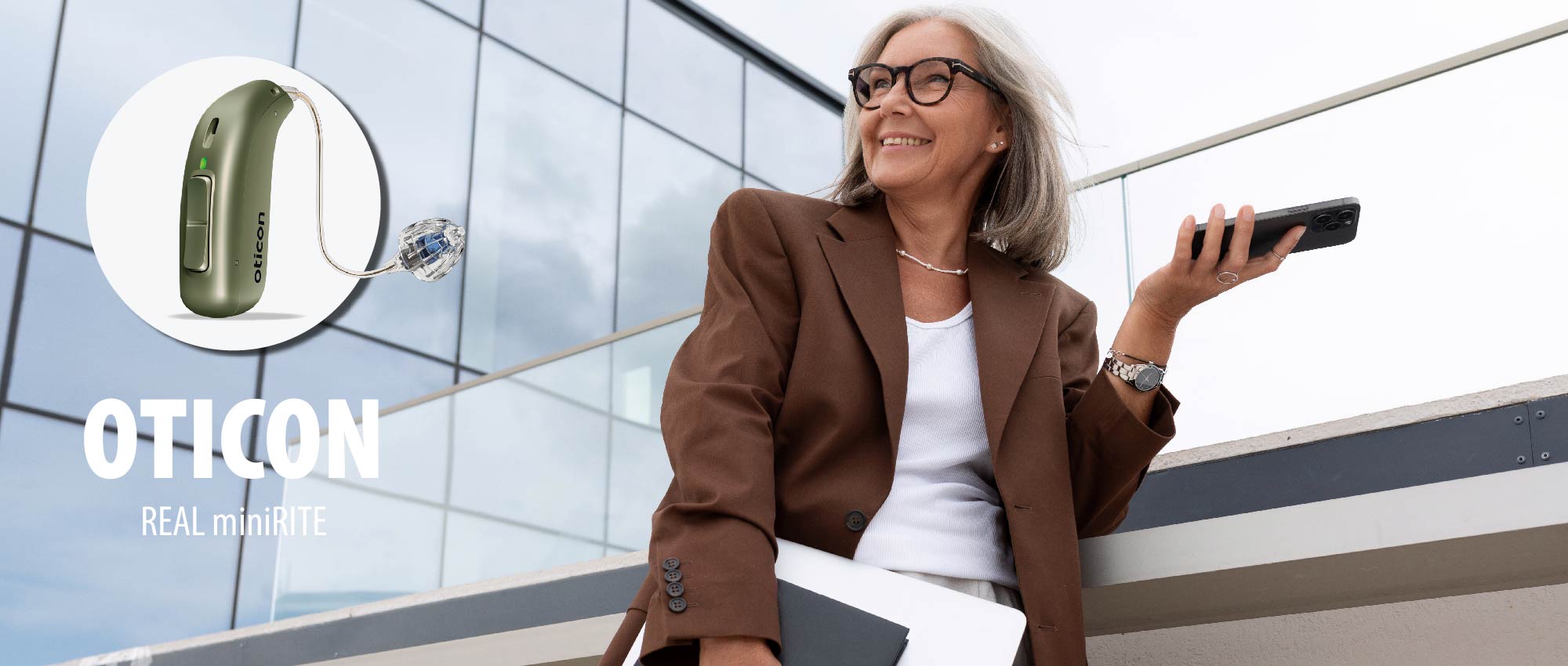 Ältere Frau mit Bluetooth Hörgerät hält Smartphone und Laptop vor moderner Glasfassade, zeigt Alltag mit Hörgerät​​​​​​​​​​​​​​​​​​​​​​​​​​​​​​​​