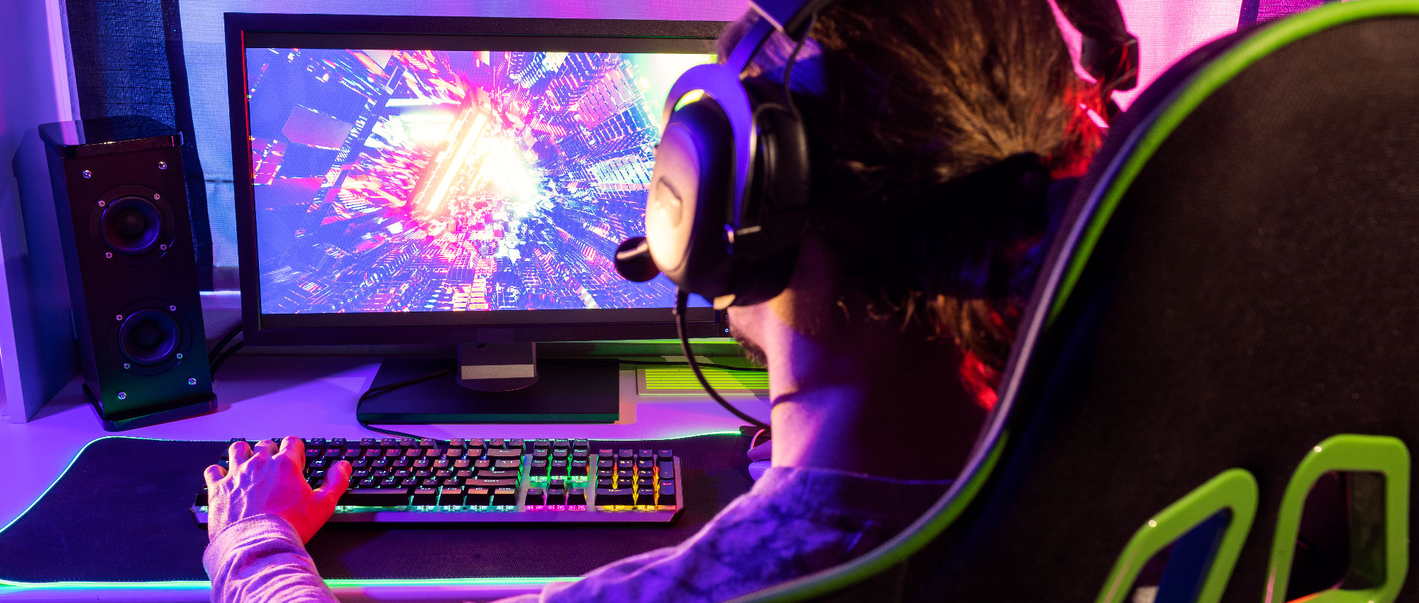 Mann mit Gaming-Kopfhörer spielt am PC mit RGB-Tastatur und Lautsprecher in bunter Gaming-Umgebung.​​​​​​​​​​​​​​​​​​​​​​​​​​​​​​​​​​​​​​​​​​​​​​