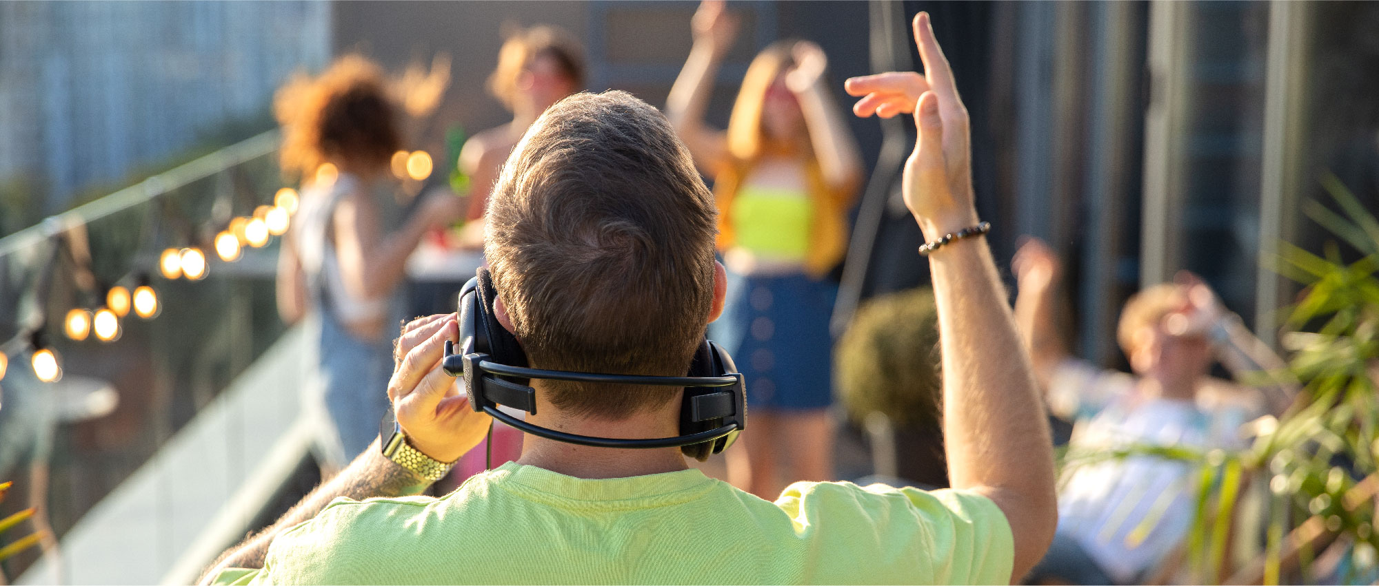 DJ mit Kopfhörern legt Musik auf Balkon, Gäste tanzen und feiern bei sonnigem Wetter mit Lichterkette im Hintergrund​​​​​​​​​​​​​​​​​​​​​​​​​​​​​