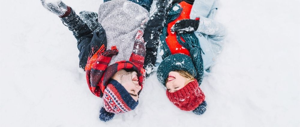 Zwei Personen liegen im Schnee, tragen warme Mützen, Schals und Jacken, genießen den Wintertag draußen im Schnee.​​​​​​​​​​​​​​​​​​​​​​​​​​​​​​​​