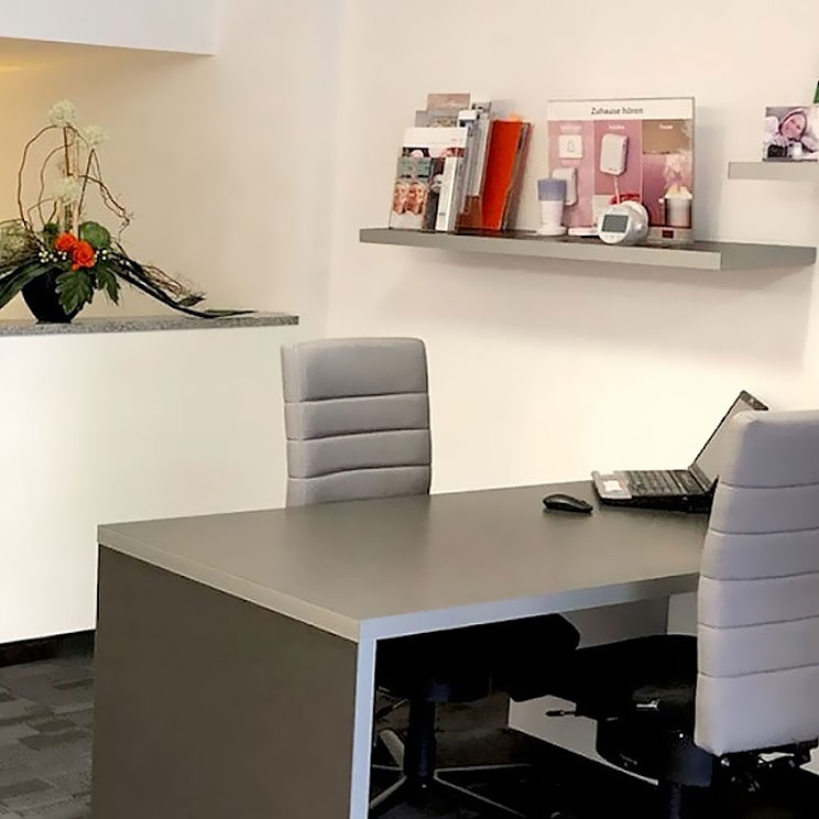 Moderner Beratungsraum mit grauem Schreibtisch, zwei Stühlen und Informationsmaterial zu Hörgeräten an der Wand.​​​​​​​​​​​​​​​​​​​​​​​​​​​​​​​​​