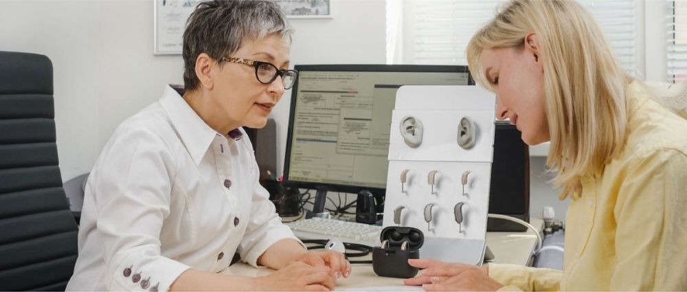 Fachfrau berät Kundin zu verschiedenen Hörgeräten in heller Praxis mit Computer und Hörgeräte-Modellen​​​​​​​​​​​​​​​​​​​​​​​​​​​​​​​​​​​​​​​​​​​