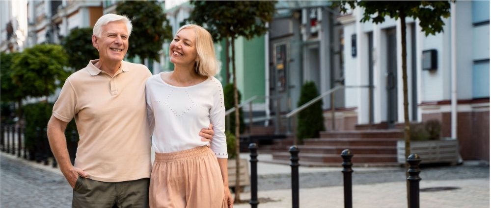 Älteres Paar geht Arm in Arm durch eine ruhige Stadtstraße mit Häusern und Bäumen im Hintergrund​​​​​​​​​​​​​​​​​​​​​​​​​​​​​​​​​​​​​​​​​​​​​​​​​