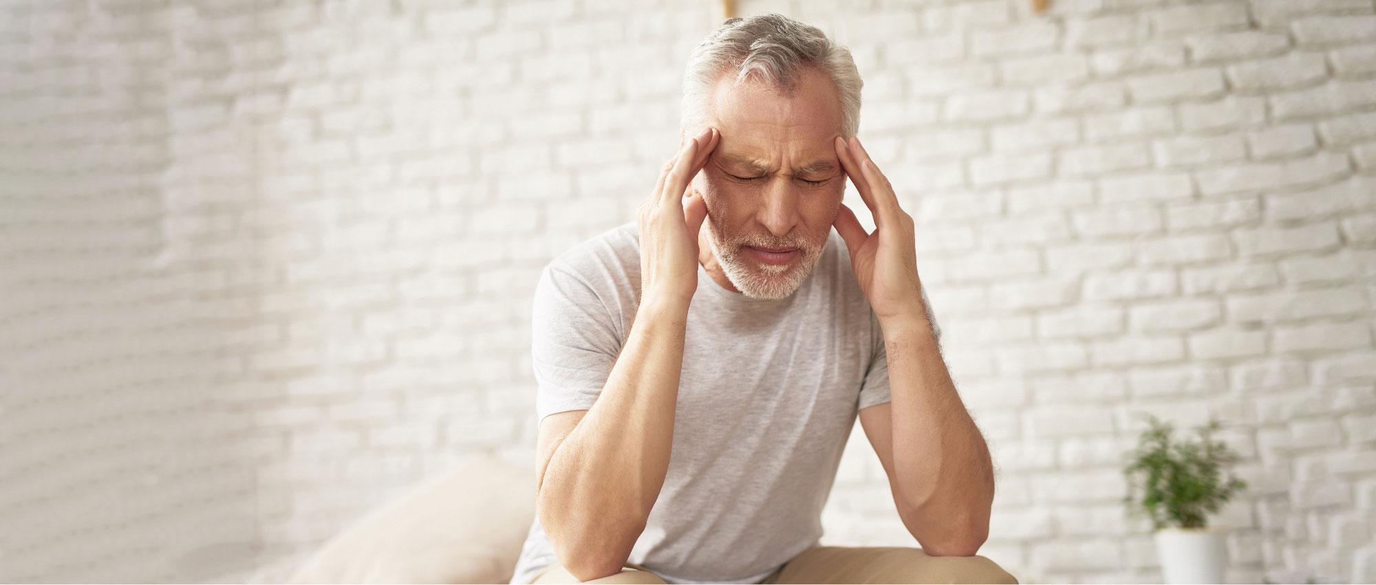 Älterer Mann sitzt vor weißer Wand und hält sich mit geschlossenen Augen die Schläfen bei Kopfschmerzen​​​​​​​​​​​​​​​​​​​​​​​​​​​​​​​​​​​​​​​​​​