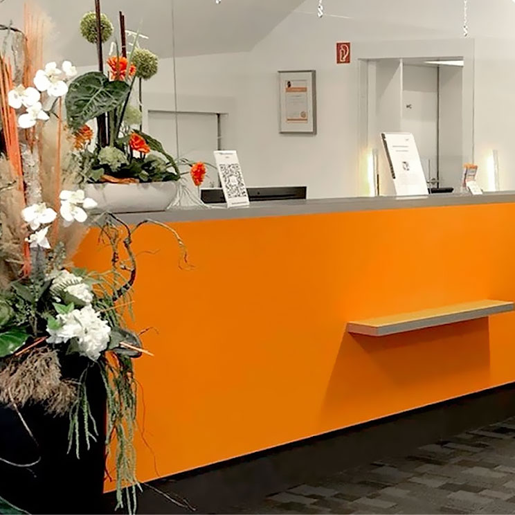 Moderner Empfang mit orangefarbenem Tresen und Blumendekoration in heller Praxisumgebung für Hörgeräte.​​​​​​​​​​​​​​​​​​​​​​​​​​​​​​​​​​​​​​​​​​