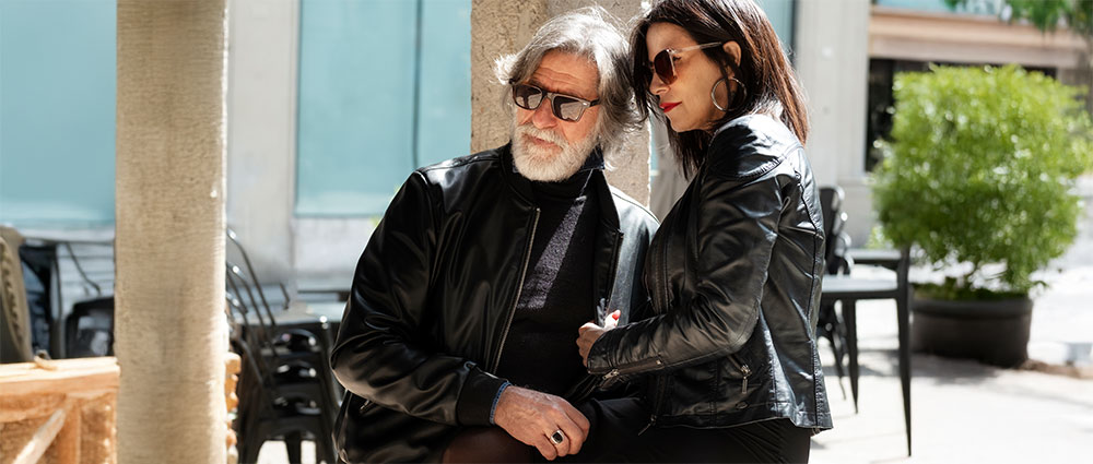 Älterer Mann und Frau tragen Sonnenbrillen und Lederjacken, sitzen draußen bei sonnigem Wetter auf einer Bank.​​​​​​​​​​​​​​​​​​​​​​​​​​​​​​​​​​​