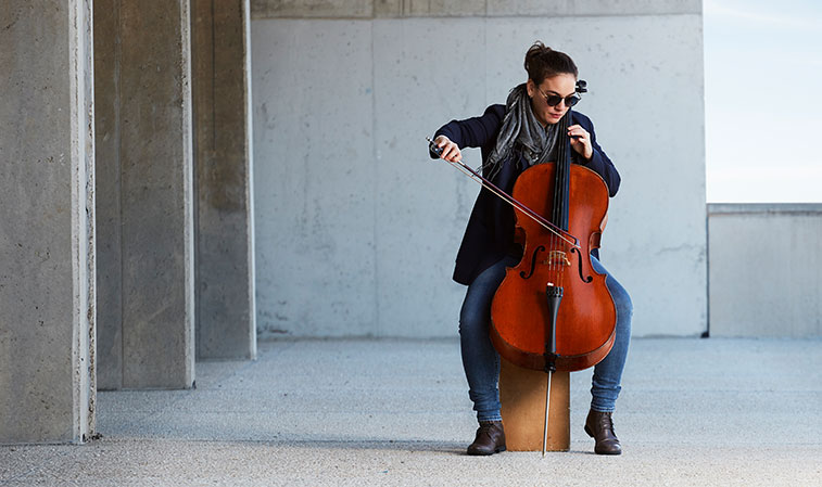 Junge Frau spielt Cello mit In-Ear-Monitoring vor Betonwand in moderner, ruhiger Umgebung​​​​​​​​​​​​​​​​​​​​​​​​​​​​​​​​​​​​​​​​​​​​​​​​​​​​​​​​