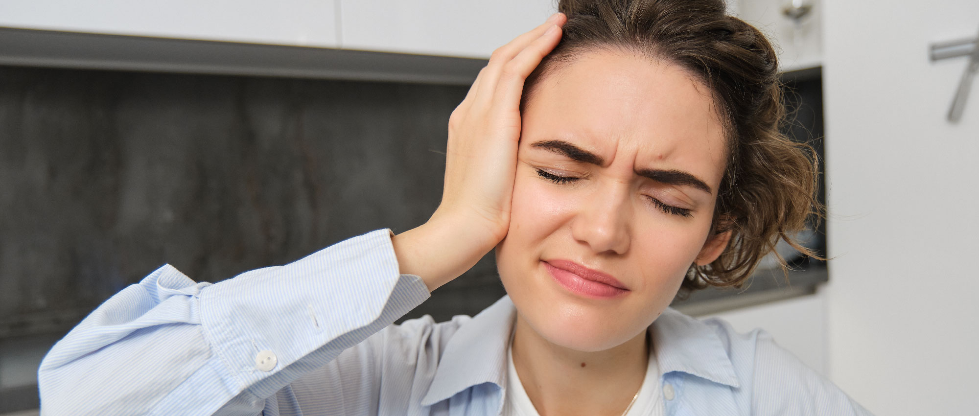 Junge Frau im hellen Hemd fasst sich schmerzhaft ans Ohr in moderner Küche, zeigt Symptome von Innenohr-Schwindel.​​​​​​​​​​​​​​​​​​​​​​​​​​​​​​​