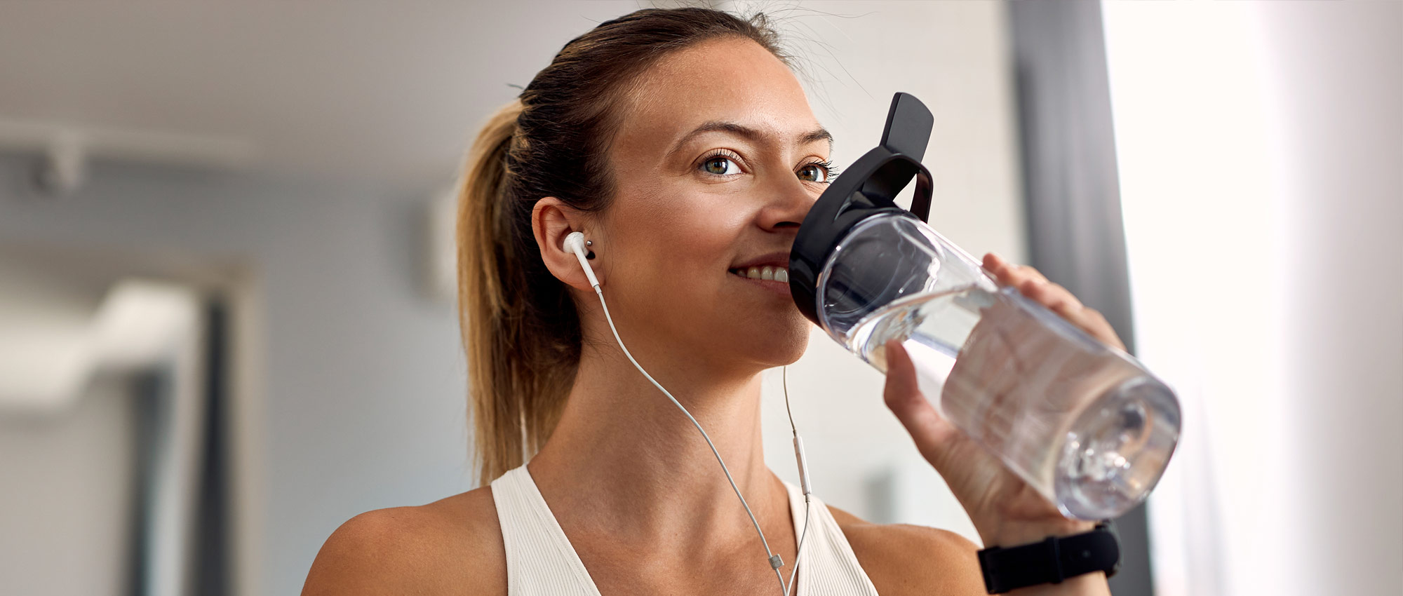 Frau mit weißen Kopfhörern trinkt Wasser aus Trinkflasche in heller Innenraumumgebung nach dem Sport.​​​​​​​​​​​​​​​​​​​​​​​​​​​​​​​​​​​​​​​​​​​​