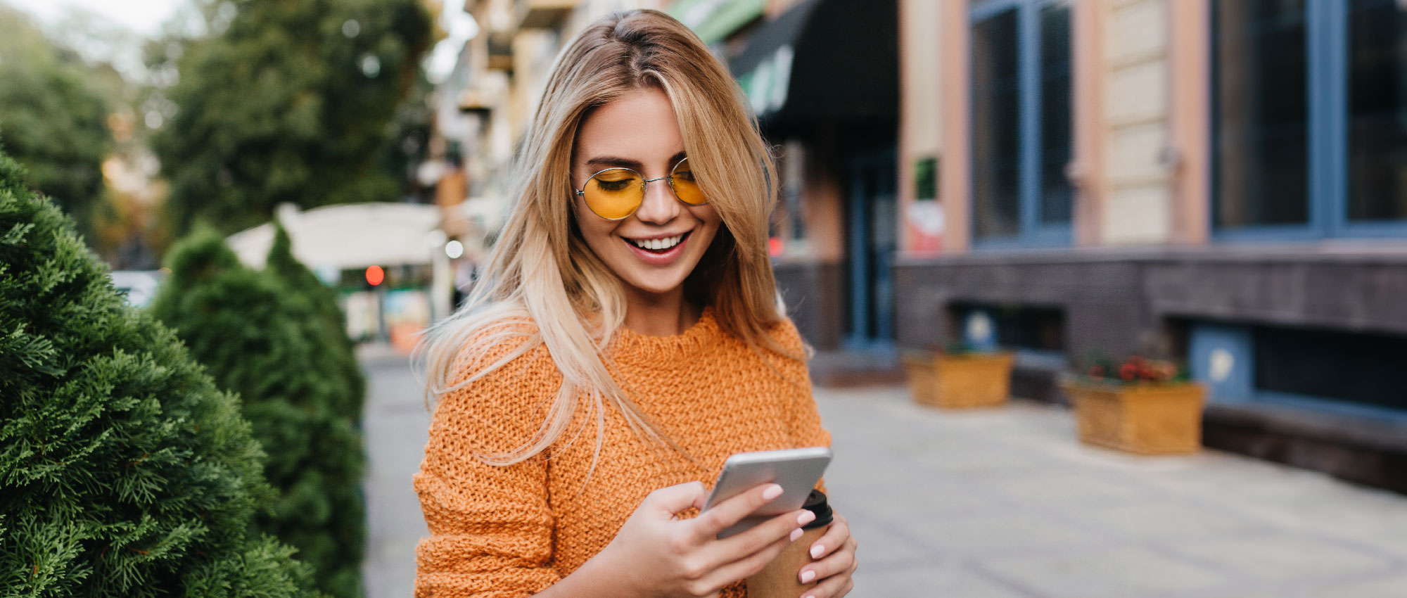 Junge Frau mit orangenem Pullover und gelber Sonnenbrille nutzt Smartphone auf Straße mit Pflanzen im Hintergrund​​​​​​​​​​​​​​​​​​​​​​​​​​​​​​​​