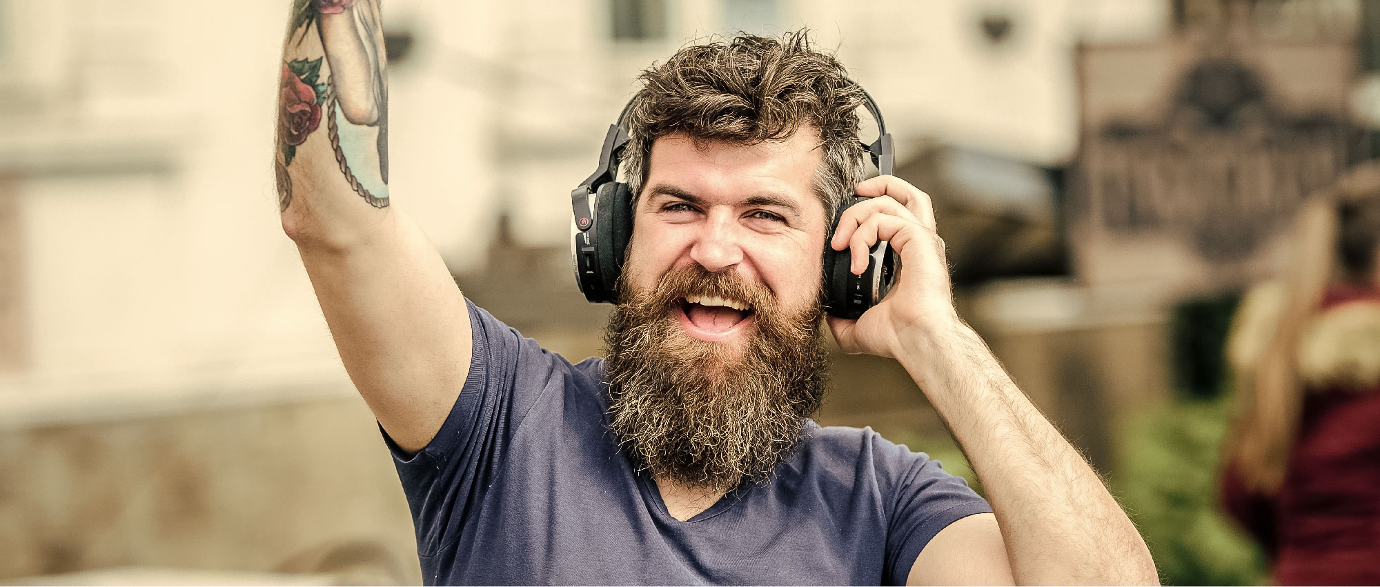 Fröhlicher Mann mit Bart und Tattoo trägt Kopfhörer und genießt Heavy Metal Musik im urbanen Umfeld.​​​​​​​​​​​​​​​​​​​​​​​​​​​​​​​​​​​​​​​​​​​​​