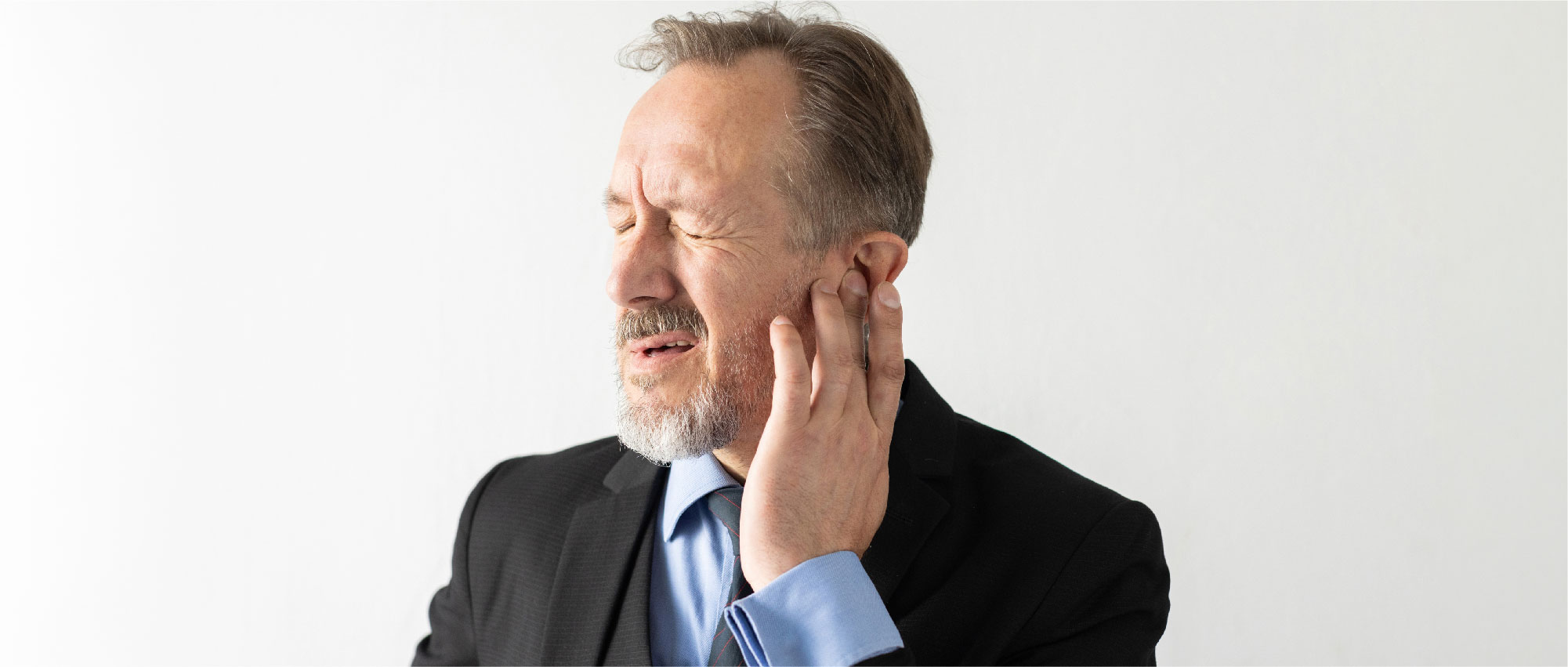 Geschäftsmann im Anzug fasst sich schmerzverzerrt ans Ohr vor weißem Hintergrund, Symbol für Ohrenschmerzen und Ohrenpflege​​​​​​​​​​​​​​​​​​​​​​