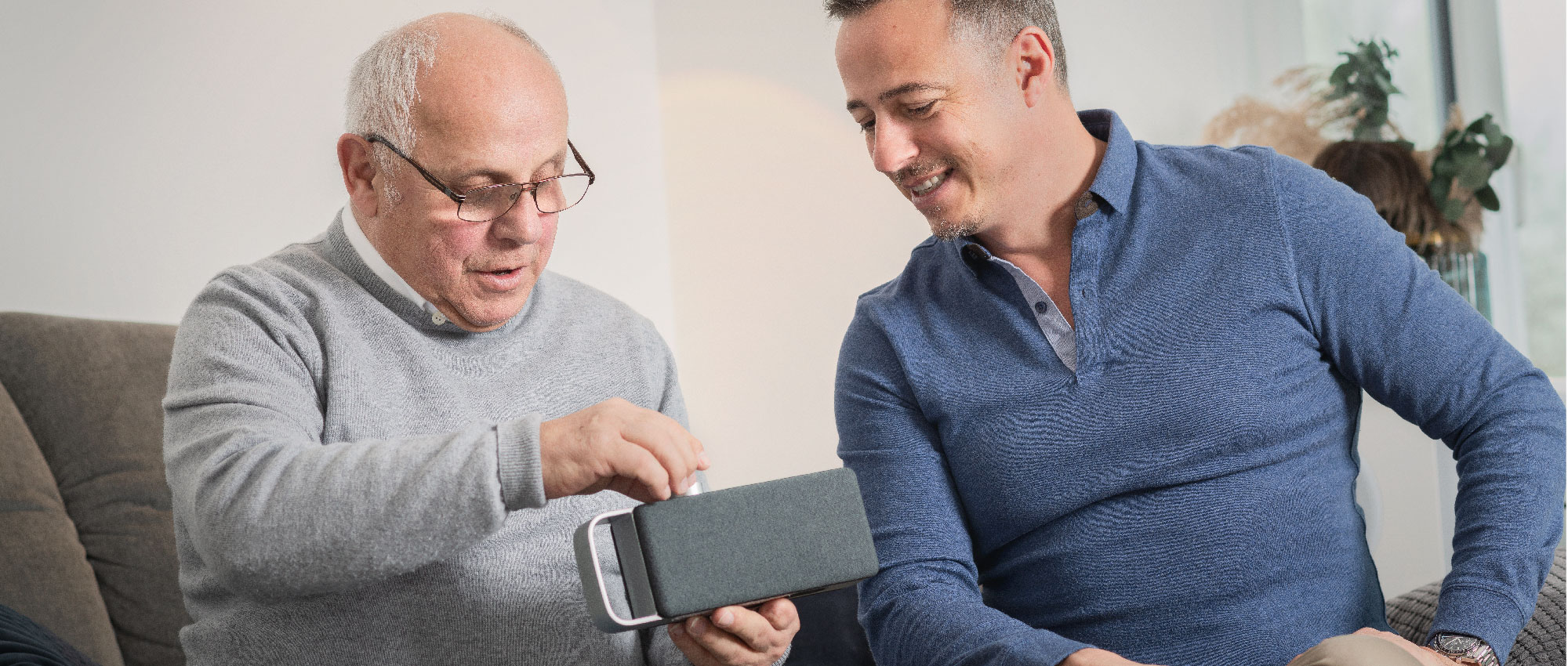 Zwei Männer sitzen im Wohnzimmer und testen einen tragbaren Lautsprecher. Die Stimmung wirkt entspannt und freundlich.​​​​​​​​​​​​​​​​​​​​​​​​​​​