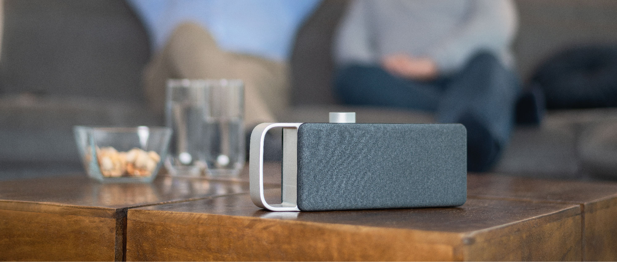 Moderner Lautsprecher steht auf Holztisch mit Gläsern und Snacks, im Hintergrund Sofa mit Personen im Wohnzimmer.​​​​​​​​​​​​​​​​​​​​​​​​​​​​​​​​
