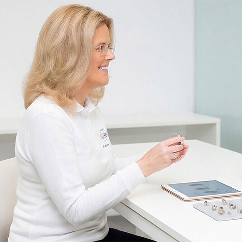 Frau in weißem Pullover passt Hörgerät an Tisch mit Tablet und Hörgerätezubehör an, helle Praxis​​​​​​​​​​​​​​​​​​​​​​​​​​​​​​​​​​​​​​​​​​​​​​​​​