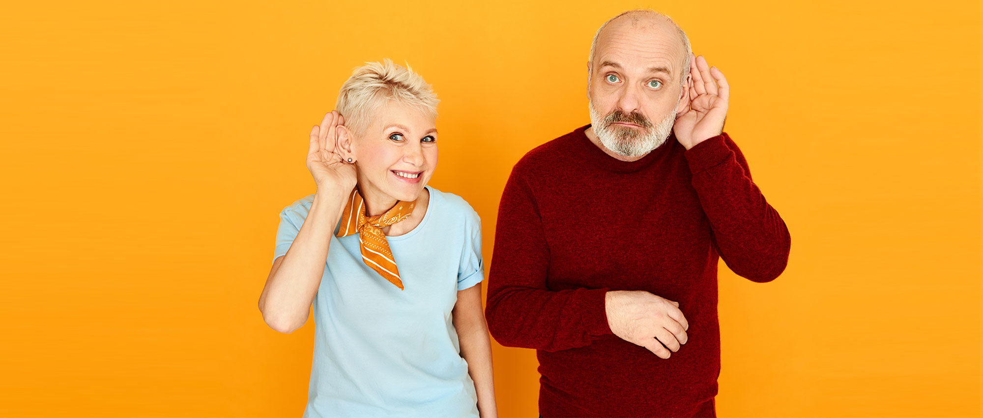 Ein älteres Paar hält sich die Hände ans Ohr vor orangefarbenem Hintergrund und symbolisiert Hörprobleme und Hörverständnis.​​​​​​​​​​​​​​​​​​​​​