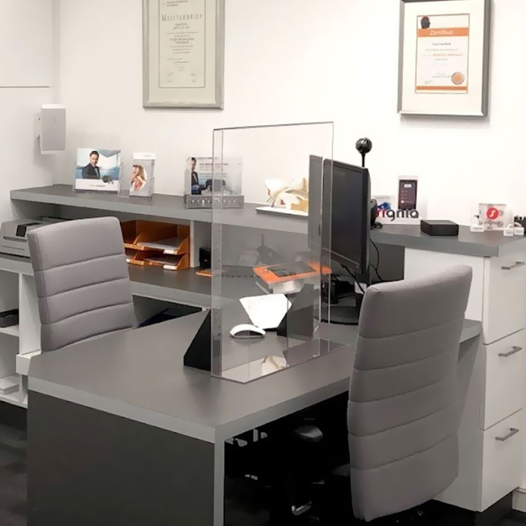 Moderner Beratungsraum mit zwei grauen Stühlen, Schreibtisch, Hygieneschutz und Computer im Hörgerätezentrum Waldsassen.​​​​​​​​​​​​​​​​​​​​​​​​​